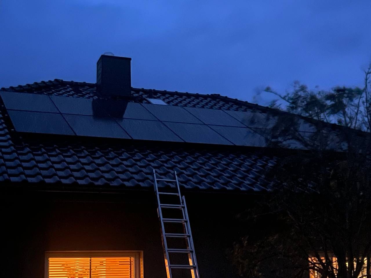 Panel słoneczny na dachu domu z drabina opartą o ścianę, widok wieczorową porą, ciepłe światło z okna.