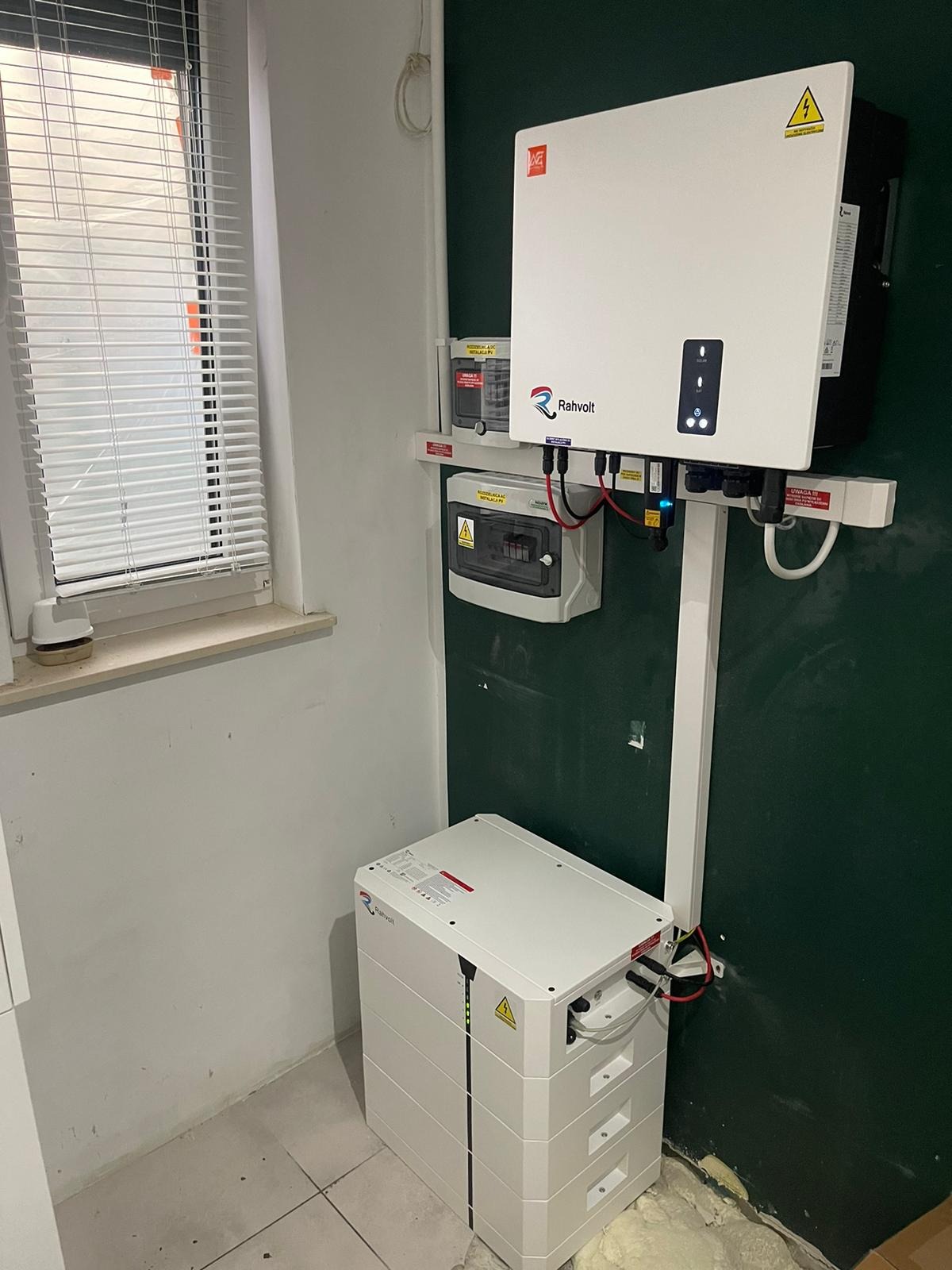 Wewnętrzna instalacja fotowoltaiczna: biała bateria akumulatorów z logo Rahvolt, podłączona do falownika i skrzynki bezpiecznikowej, zamontowane na ścianie w pomieszczeniu z oknem z żaluzjami.