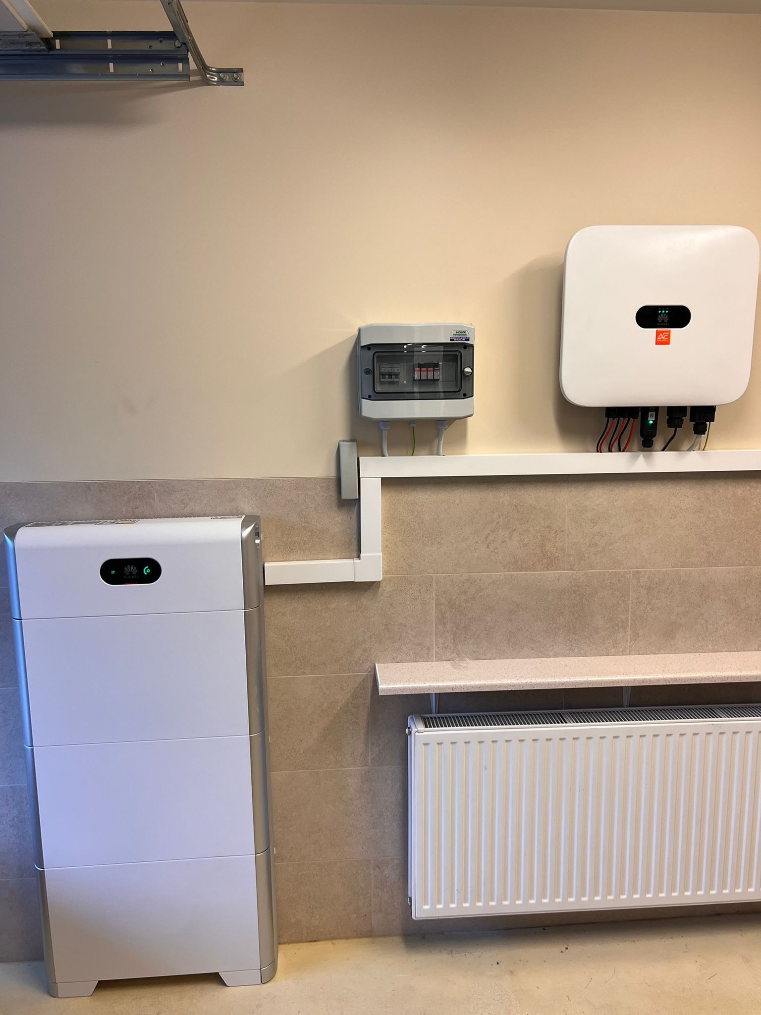 Biała bateria magazynu energii obok grzejnika i rozdzielnicy elektrycznej, z białym inwerterem fotowoltaicznym zamontowanym na ścianie powyżej, w pomieszczeniu z beżowymi ścianami i płytkami.