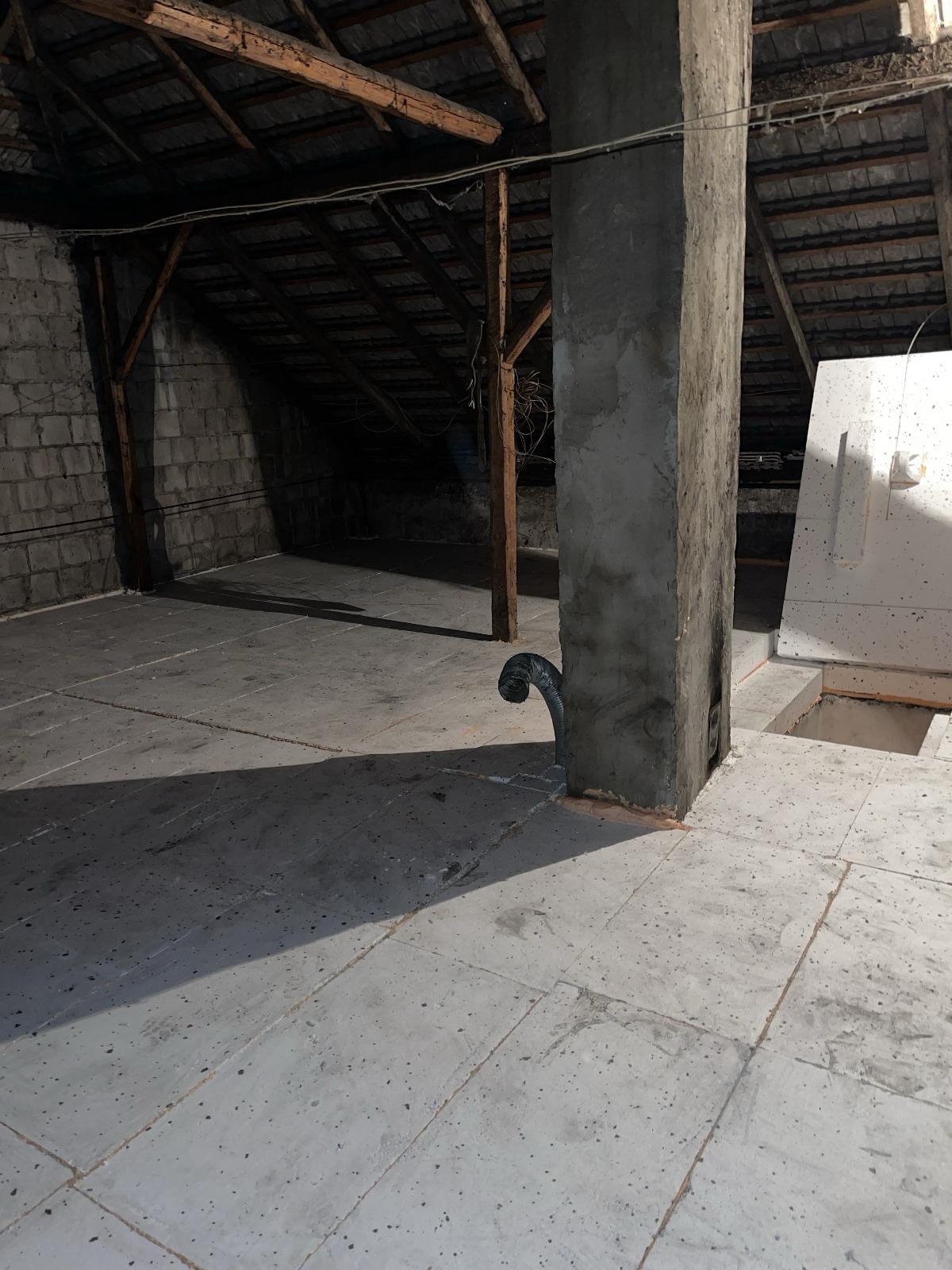 Poddasze po wylewce, widoczny komin obłożony betonem z wystającą rurą wentylacyjną, surowa cegła na ścianach bocznych i drewniana konstrukcja dachu.