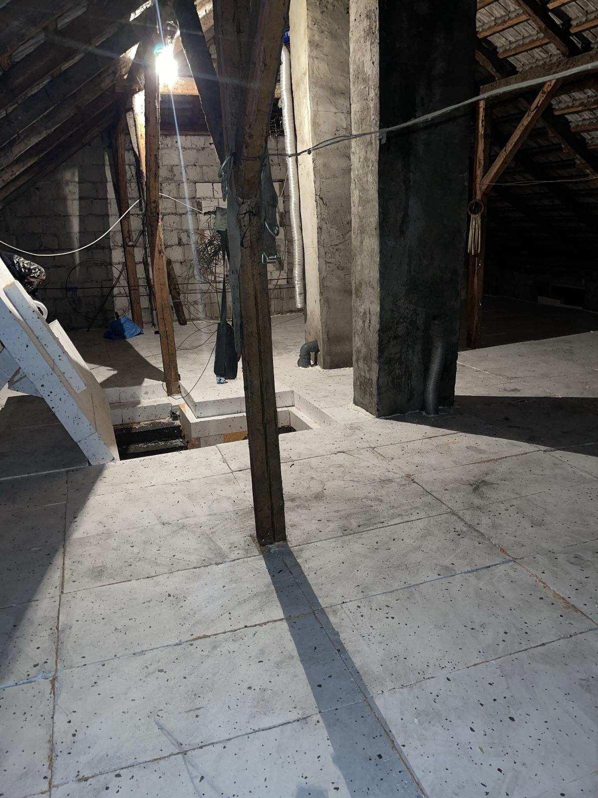 Poddasze ze starą więźbą dachową i betonową podłogą, widoczne otwory w podłodze i przygotowane płyty do montażu, słabe oświetlenie