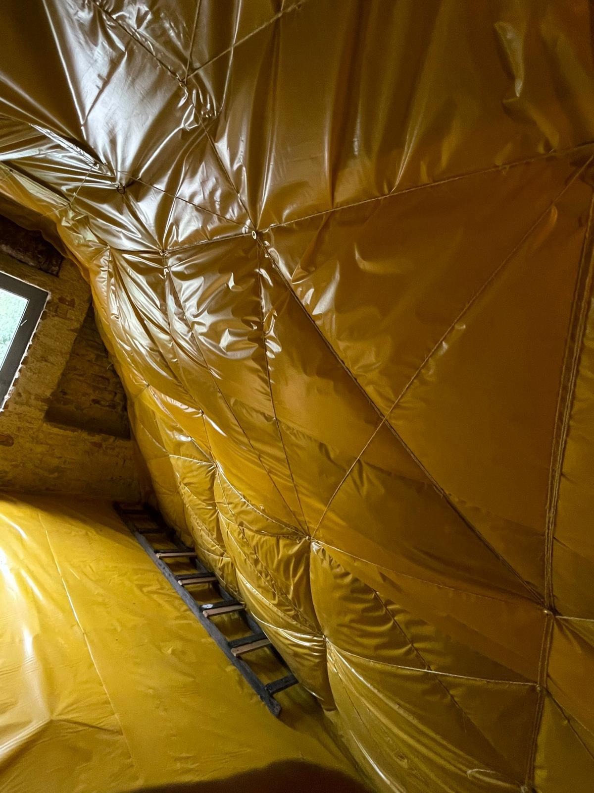 Poddasze w trakcie ocieplania, pokryte żółtą folią z widocznymi szwami i mocowaniami, drabina oparta o ścianę, okno w tle.