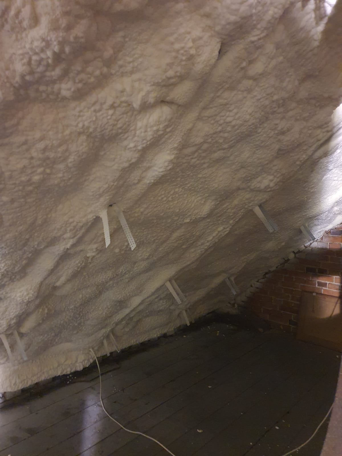 Poddasze ocieplone pianką PUR, widoczne drewniane deski podłogowe i fragment ściany z cegły, z plastikowymi elementami mocującymi piankę.