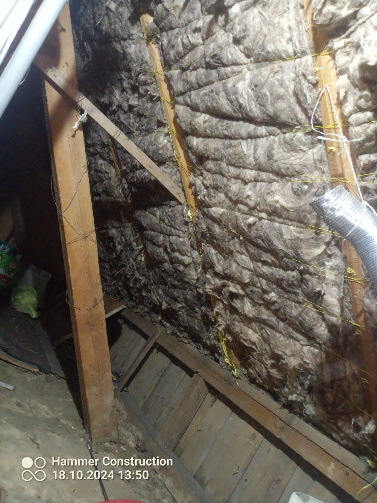 Poddasze z drewnianą konstrukcją i izolacją z wełny mineralnej, zabezpieczoną sznurkiem. Widoczny fragment rury wentylacyjnej i resztki materiałów budowlanych na podłodze.