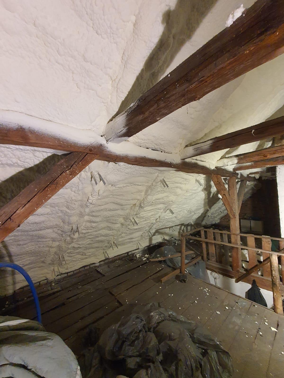 Poddasze z drewnianą konstrukcją pokryte grubą warstwą jasnej pianki izolacyjnej, widoczne metalowe elementy mocujące, drewniany pomost i worki z odpadami na podłodze.