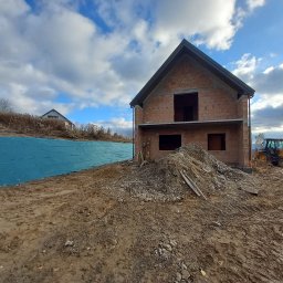 Dk-Pol - Surowy stan budowy domu z cegły, widoczny żółty traktor i sterta ziemi, błękitne niebo z chmurami w tle.