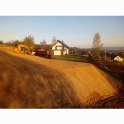 Dk-Pol - Żółta koparka i czerwona wywrotka na placu budowy w Krakowie, w tle dom jednorodzinny i drzewa.