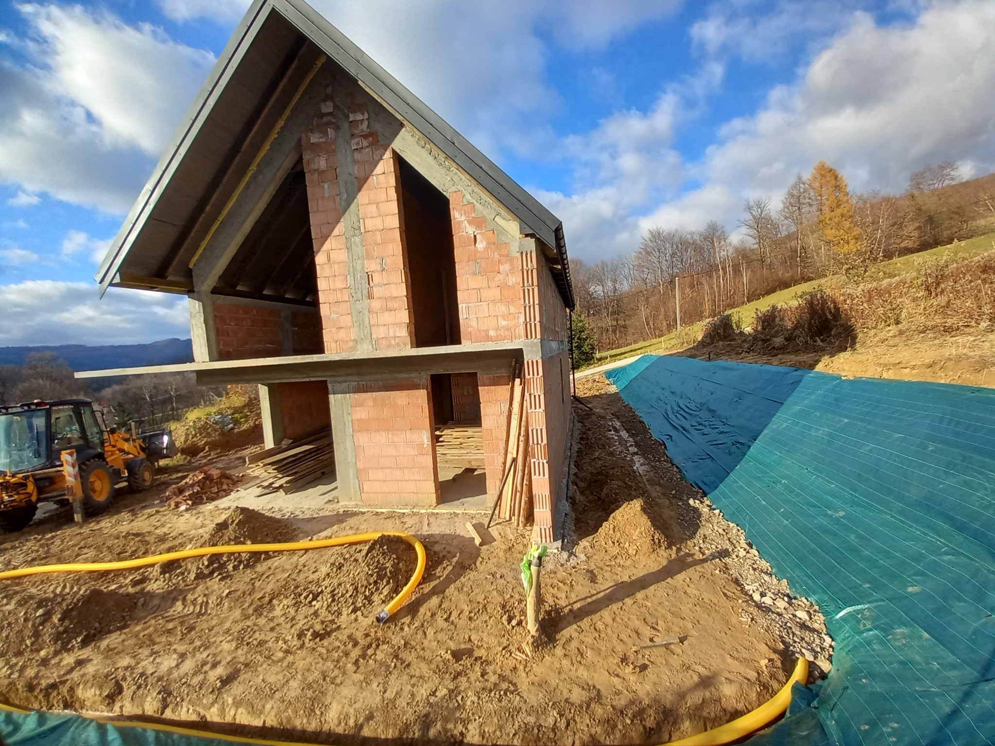 Budowa domu w stanie surowym otwartym, widoczny fundament, ściany z cegły, dach, żółty wąż i maszyna budowlana na tle ziemi i zbocza wzgórza z drzewami.