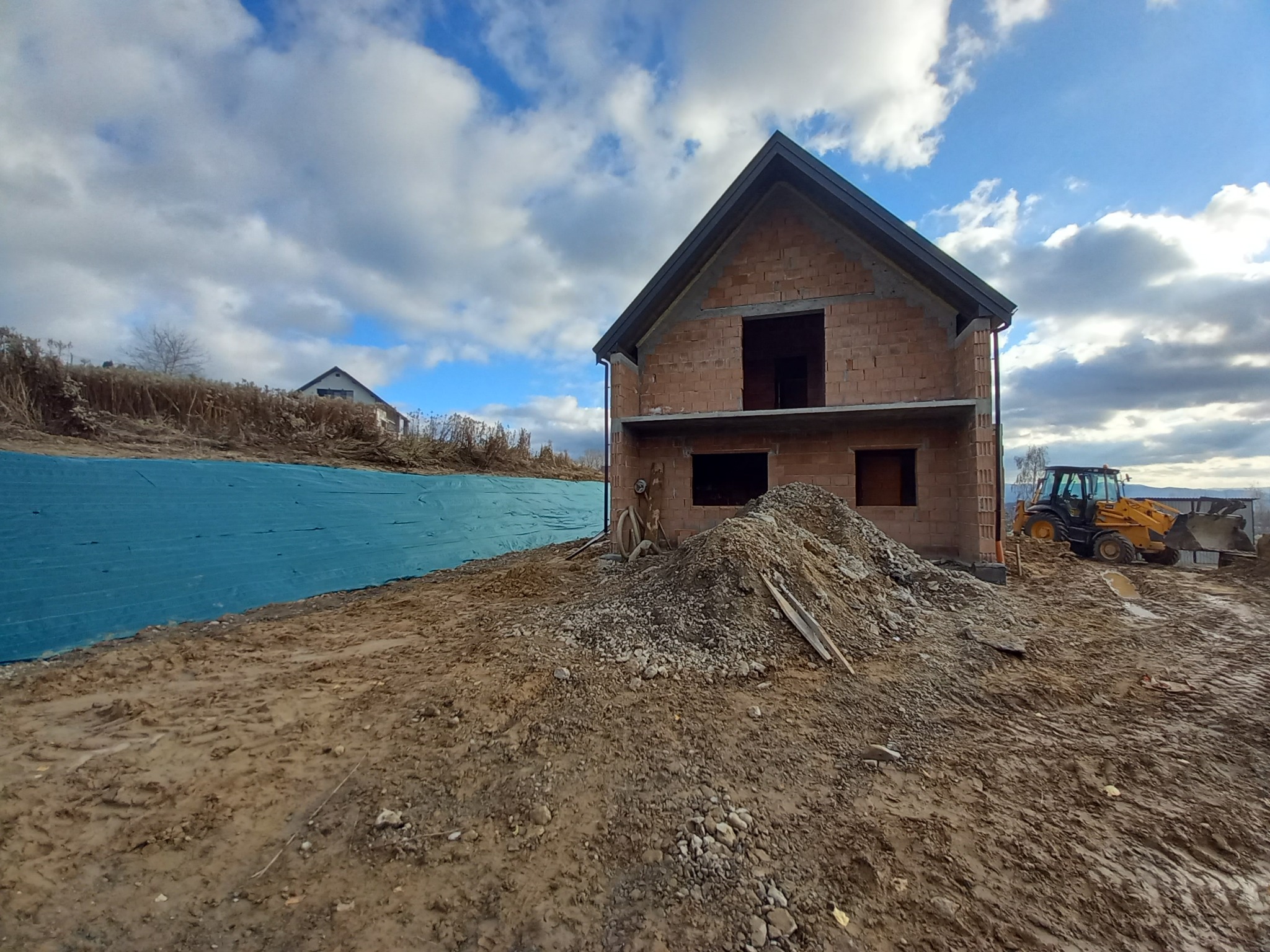 Surowy stan budowy domu z cegły, widoczny żółty traktor i sterta ziemi, błękitne niebo z chmurami w tle.