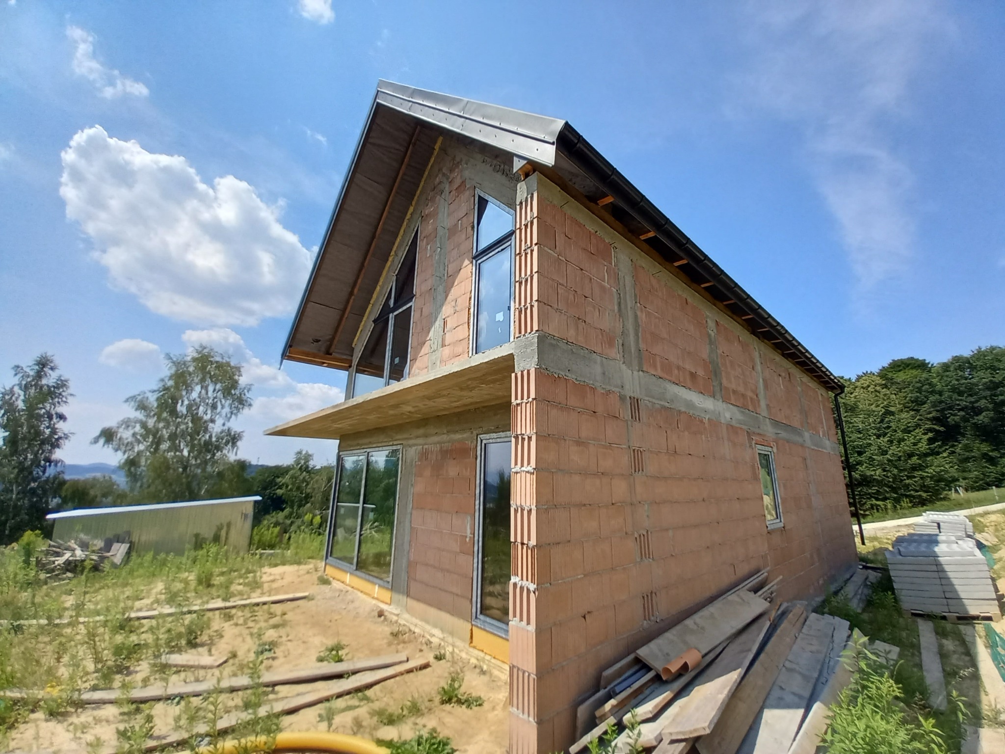 Niedokończony dom z czerwonej cegły z widocznymi oknami i elementami konstrukcyjnymi, otoczony ziemią i materiałami budowlanymi, na tle błękitnego nieba z chmurami.