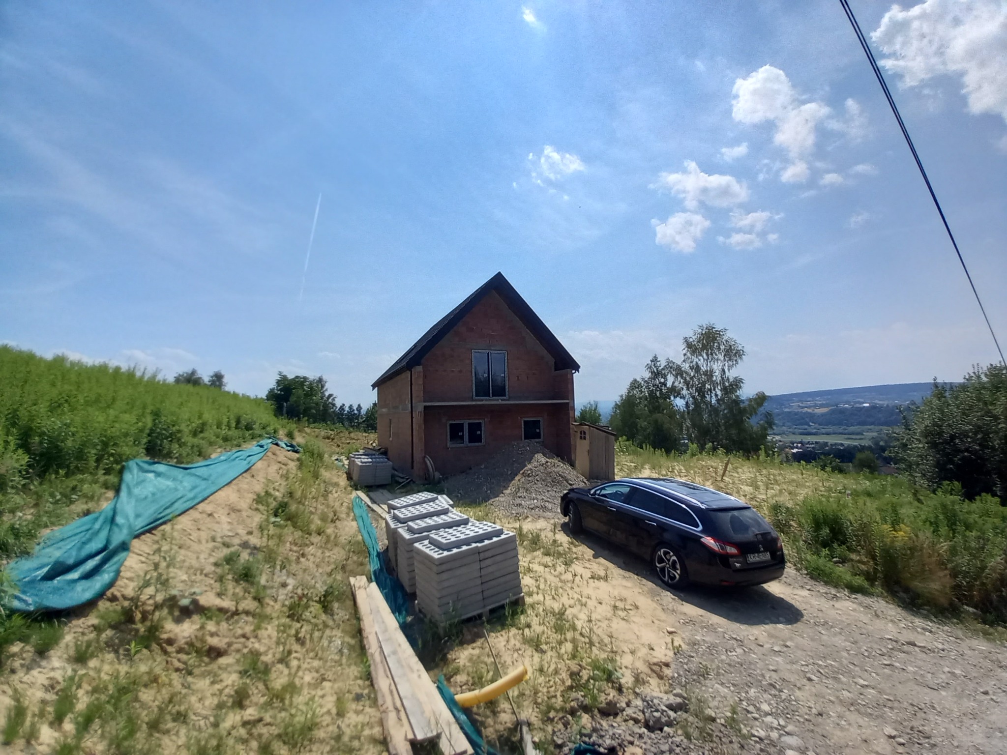 Budowa domu w trakcie: widoczny surowy stan, sterty bloczków betonowych, ziemia i samochód osobowy na działce.