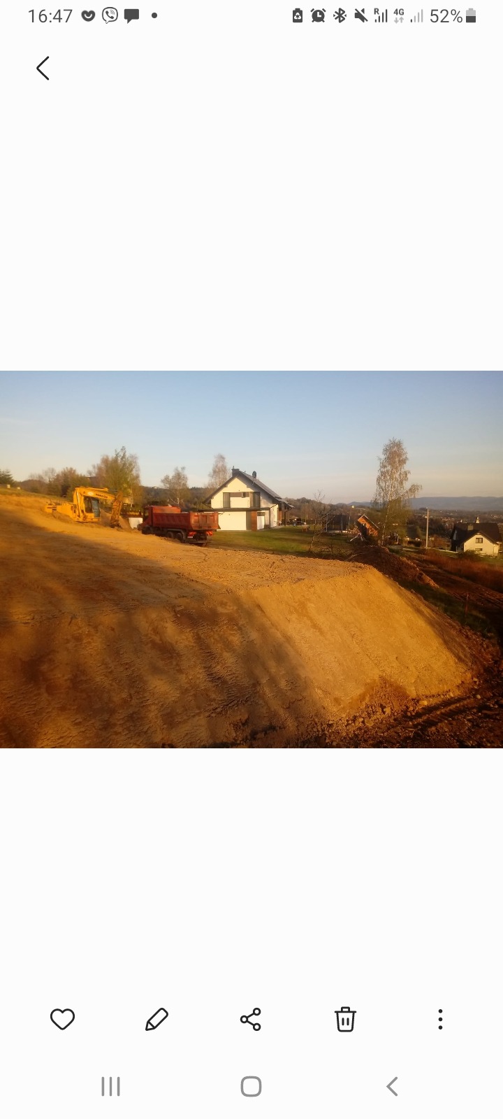 Żółta koparka i czerwona wywrotka na placu budowy w Krakowie, w tle dom jednorodzinny i drzewa.