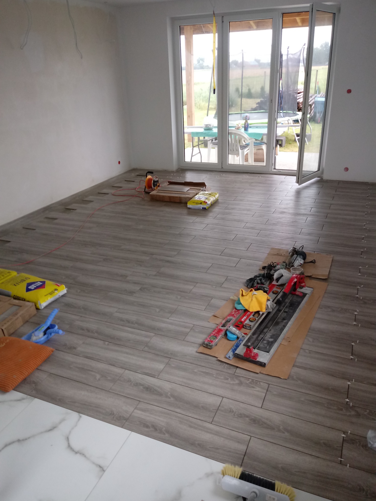 Układanie podłogi z płytek imitujących drewno w jasnym pomieszczeniu z dużym oknem tarasowym, widoczne narzędzia glazurnicze, worki z klejem i krzyżyki dystansowe, częściowo ułożona podłoga...