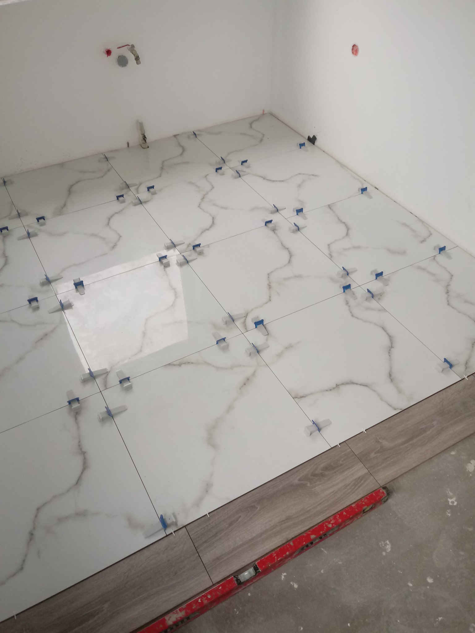 Układanie białych płytek imitujących marmur na podłodze w łazience, z użyciem niebieskich klipsów poziomujących, widoczny fragment paneli podłogowych i poziomica.