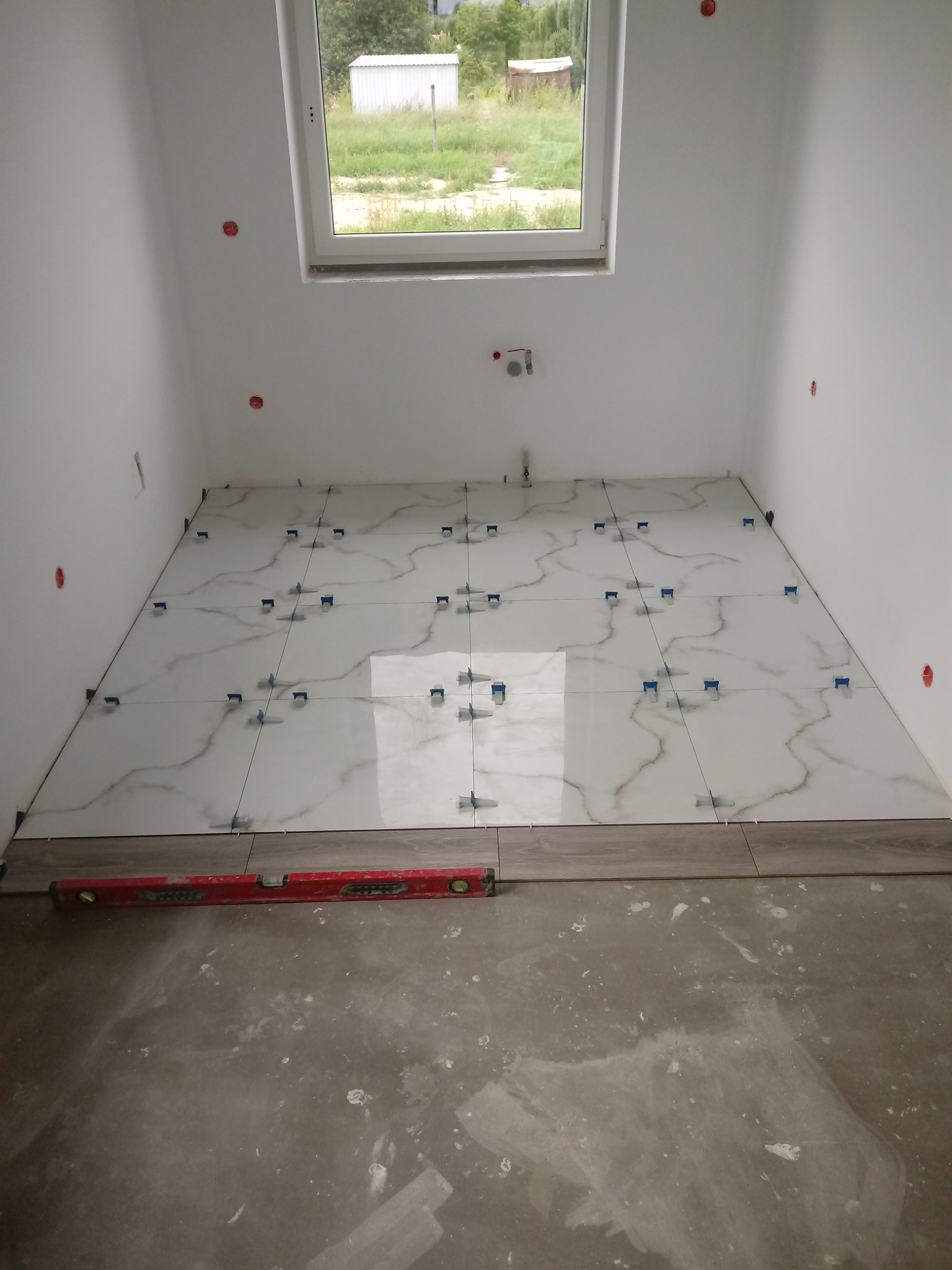 Układanie białych płytek podłogowych z szarym wzorem żyłkowania, z użyciem niebieskich klipsów poziomujących, widoczna poziomica na podłodze w trakcie prac w łazience z oknem.