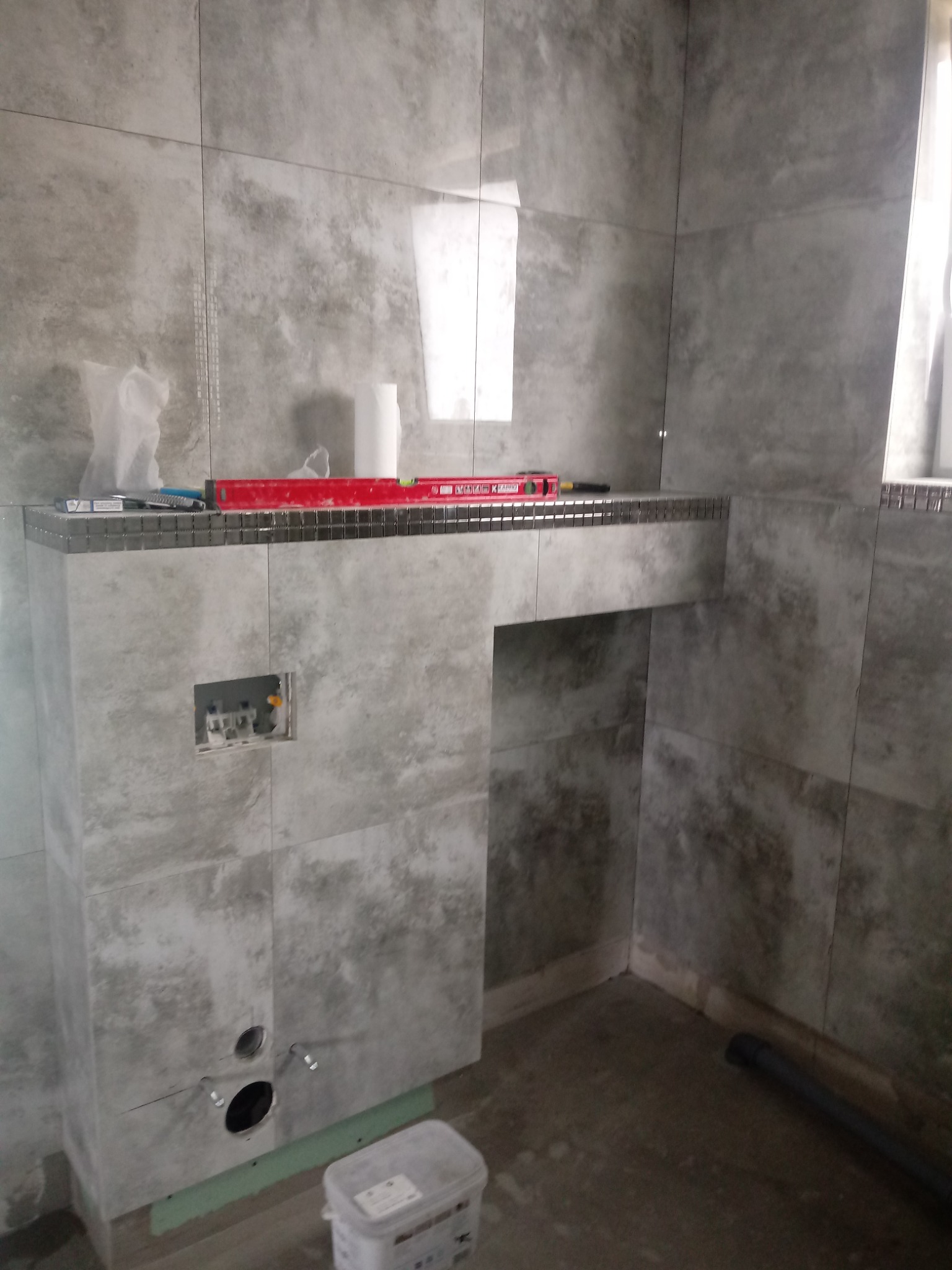 Szara łazienka w trakcie remontu, widoczne płytki imitujące beton, poziomica, puszka instalacyjna i wiadro z klejem.