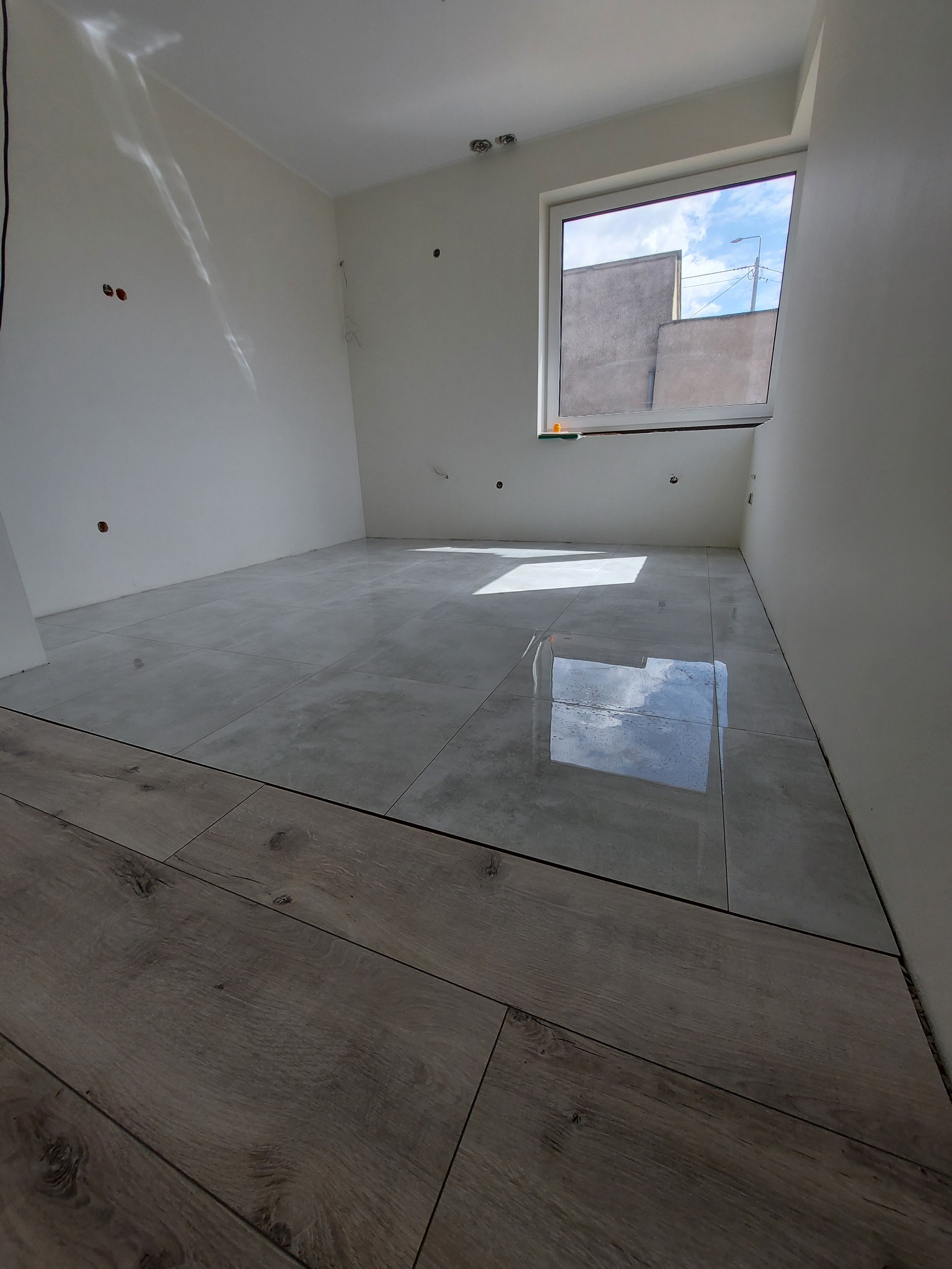 Jasnoszara, błyszcząca podłoga wyłożona płytkami w pustym pokoju, z fragmentem drewnianej podłogi w dolnej części kadru, widoczne niedokończone ściany i okno z widokiem na budynki.