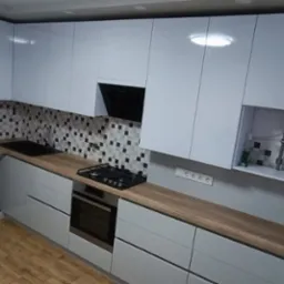 Nowoczesna kuchnia z białymi, lakierowanymi frontami, drewnianym blatem, mozaiką na ścianie, widocznym piekarnikiem, płytą gazową i lod&oacute;wką.