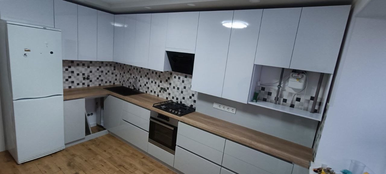 Nowoczesna kuchnia z białymi, lakierowanymi frontami, drewnianym blatem, mozaiką na ścianie, widocznym piekarnikiem, płytą gazową i lodówką.