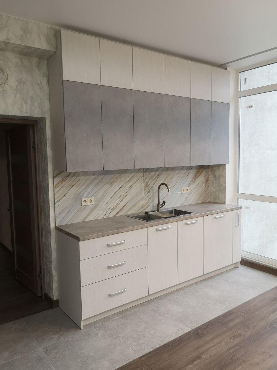Nowoczesna kuchnia z dwukolorowymi szafkami (białe i szare) i blatem imitującym kamień, z widocznym zlewem i baterią, wykończona marmurową ścianą nad blatem i szarą podłogą.