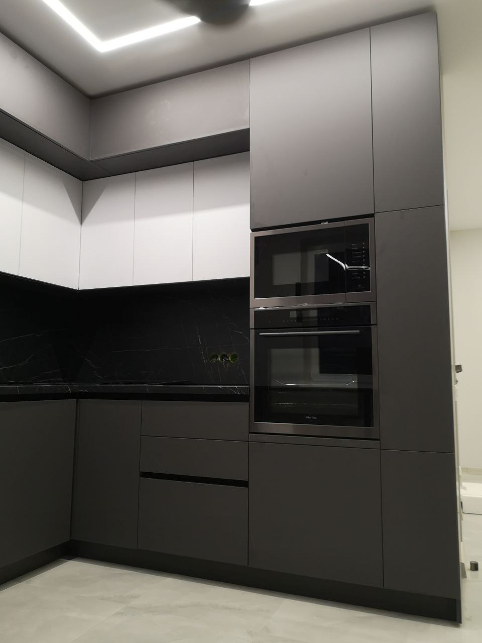 Nowoczesna kuchnia z szarymi dolnymi szafkami, białymi górnymi szafkami i czarnym blatem imitującym marmur, z wbudowanym piekarnikiem i mikrofalówką.
