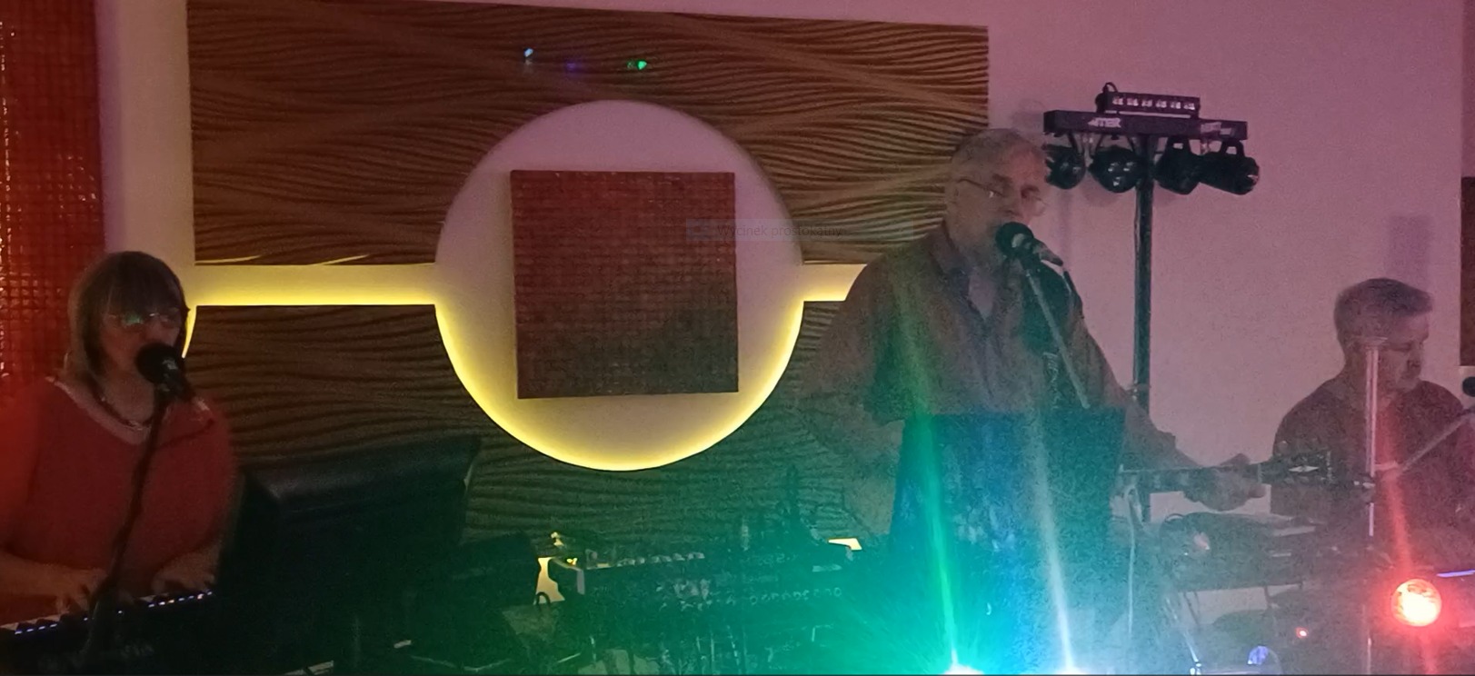 Trzyosobowy zespół grający muzykę na żywo podczas imprezy, widoczne instrumenty klawiszowe, mikrofony i oświetlenie sceniczne.