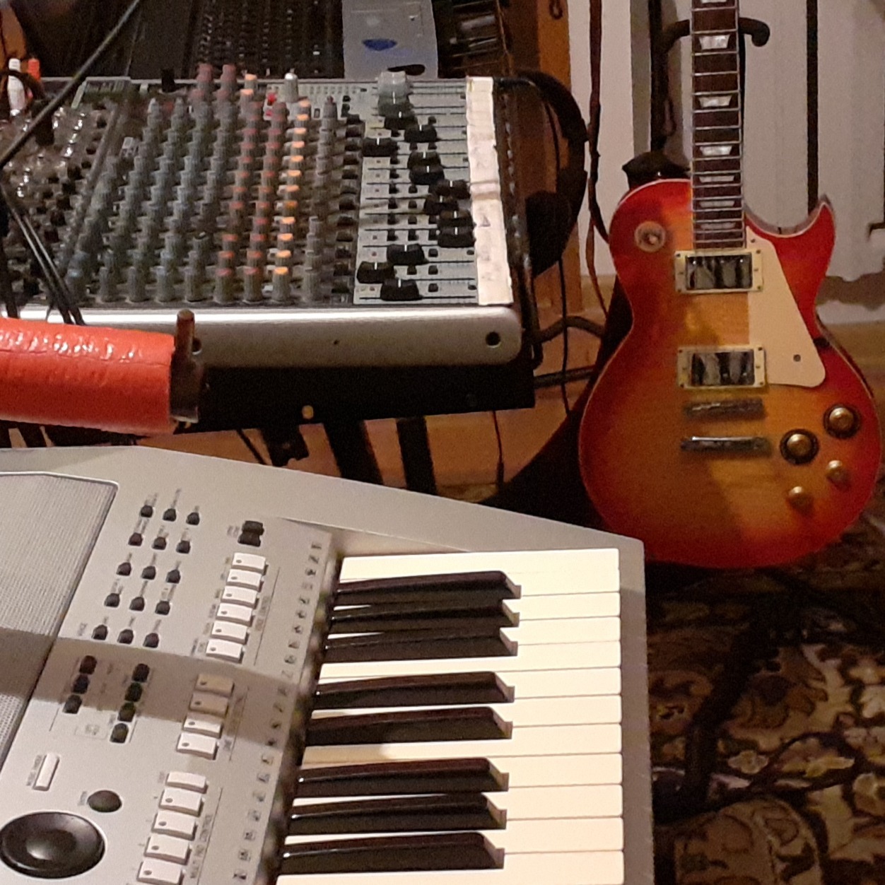 Gitara elektryczna typu Les Paul, klawisze i mikser na statywie, sprzęt muzyczny w pomieszczeniu o słabym oświetleniu.