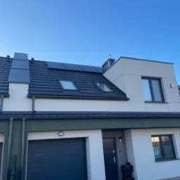 Nowoczesny dom jednorodzinny z panelami fotowoltaicznymi na dachu pokrytym ciemną dachówką, widoczne okna dachowe i komin, błękitne niebo w tle.