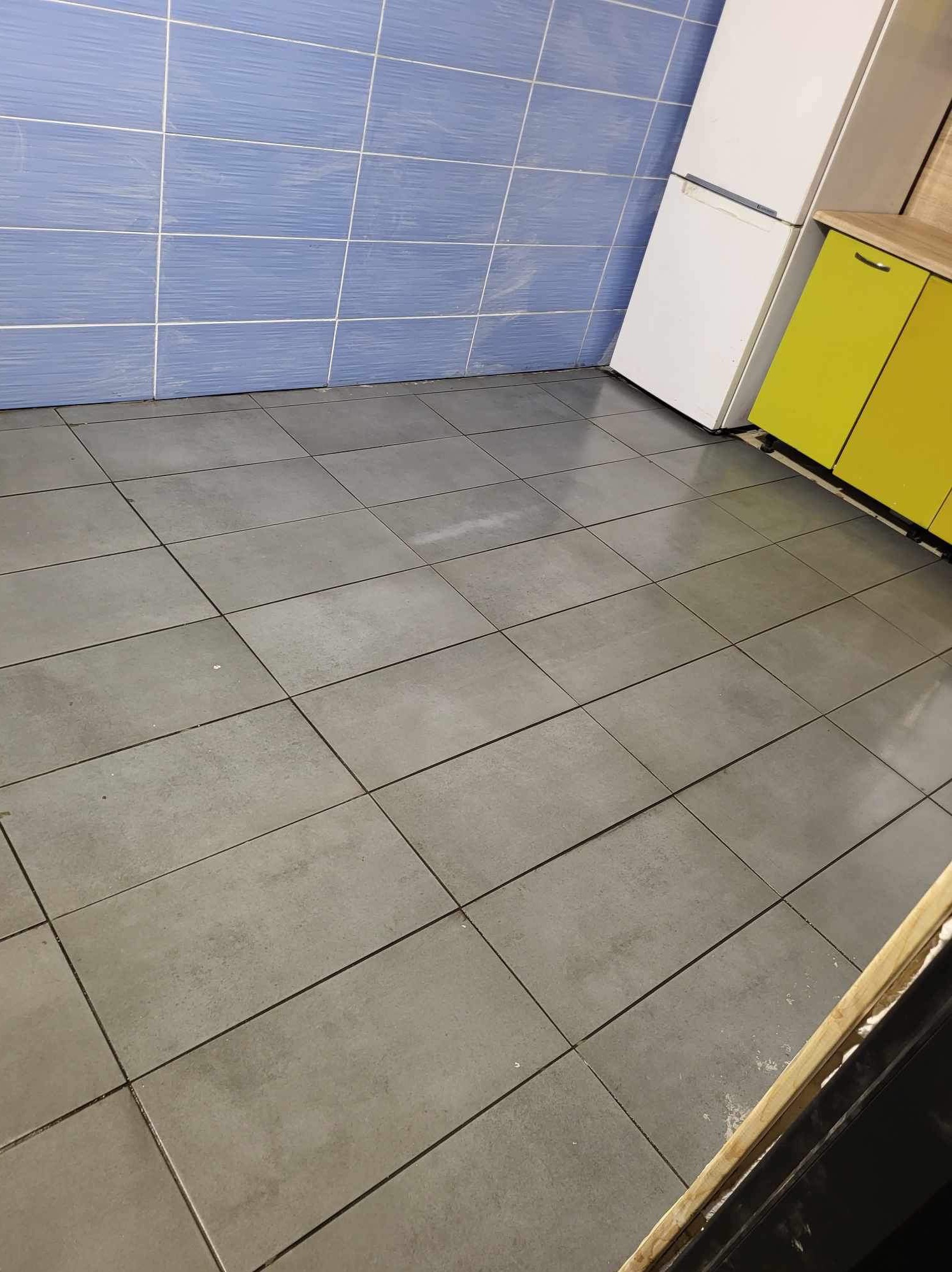 Wyłożona szarymi płytkami podłoga w kuchni z niebieskimi płytkami na ścianie i żółtymi szafkami oraz białą lodówką w rogu.