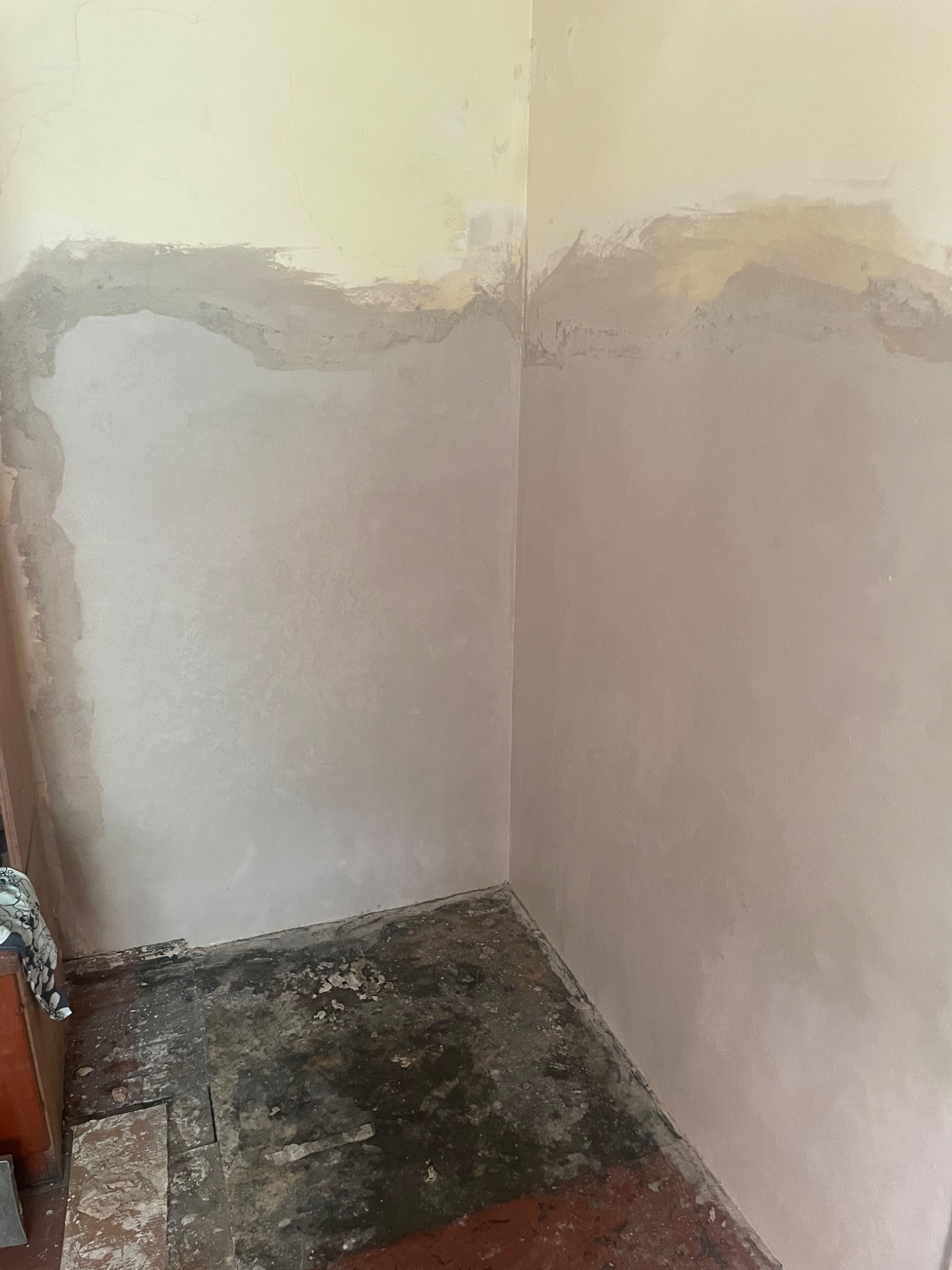 Przygotowanie ściany do malowania: gruntowanie i szpachlowanie nierówności w rogu pomieszczenia, widoczny fragment podłogi przed remontem.