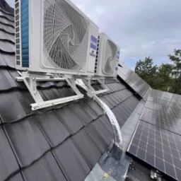 Dwa białe, zewnętrzne moduły klimatyzacji marki Haier zamontowane na metalowym stelażu na ciemnym, skośnym dachu z dachówki ceramicznej, obok paneli fotowoltaicznych, z widocznymi przewodami...