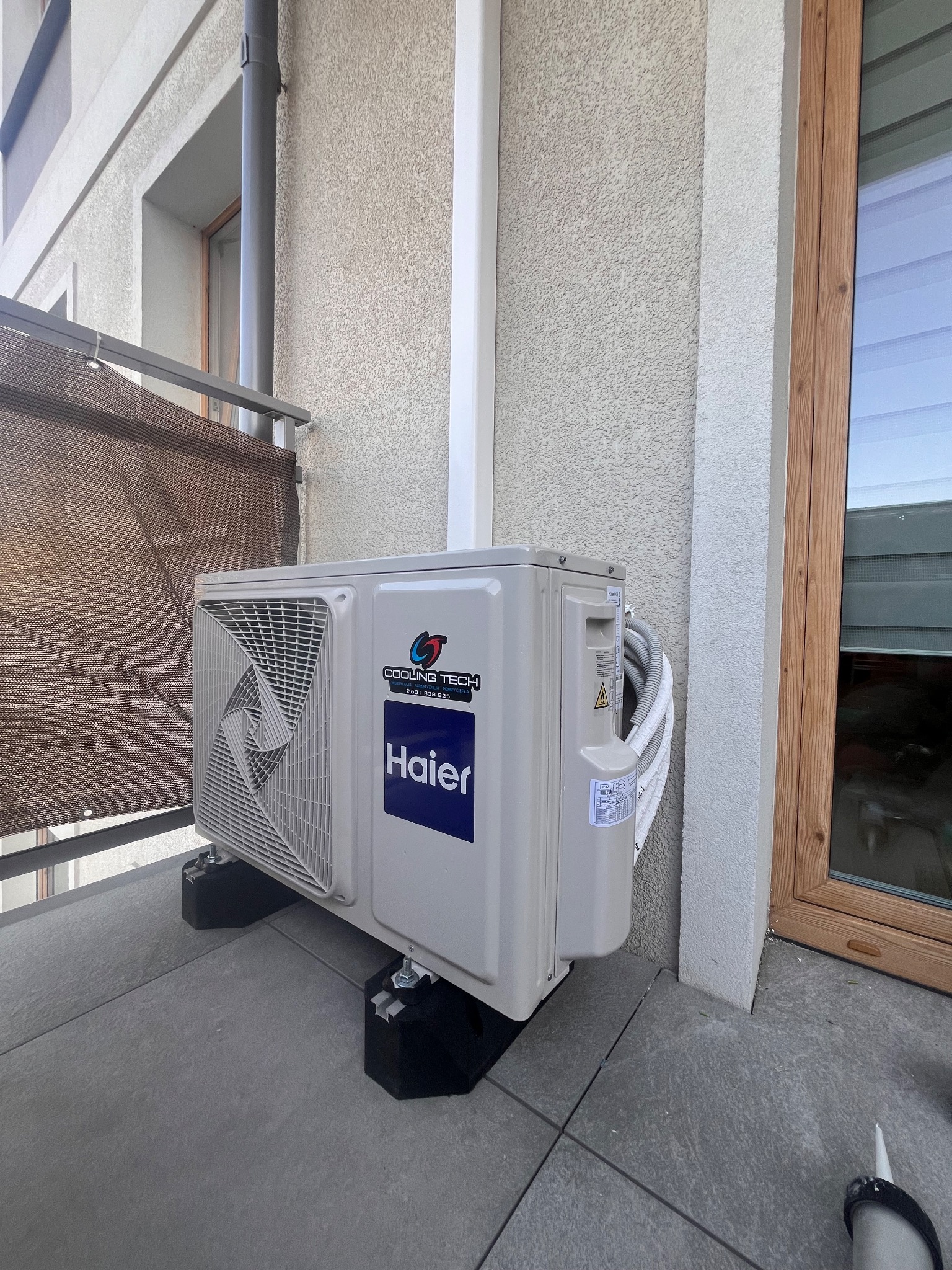 Zewnętrzna jednostka klimatyzatora Haier zamontowana na balkonie na specjalnych podkładkach tłumiących wibracje, widoczne przewody w białej osłonie i tuba silikonu uszczelniającego przy podłodze