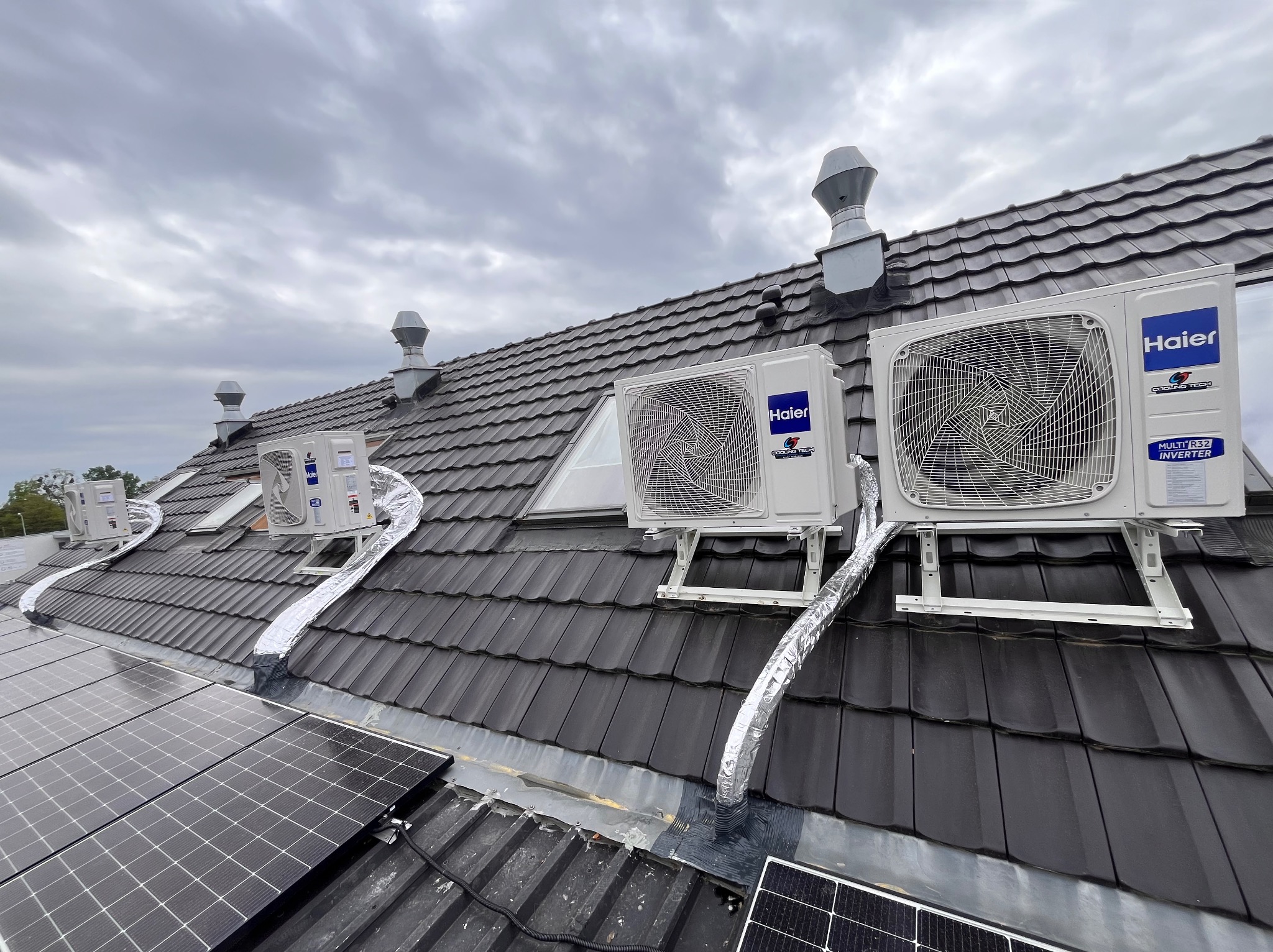 Kilka jednostek zewnętrznych klimatyzacji marki Haier zamontowanych na dachu pokrytym czarną dachówką obok paneli słonecznych, z widocznymi srebrnymi rurami odprowadzającymi.
