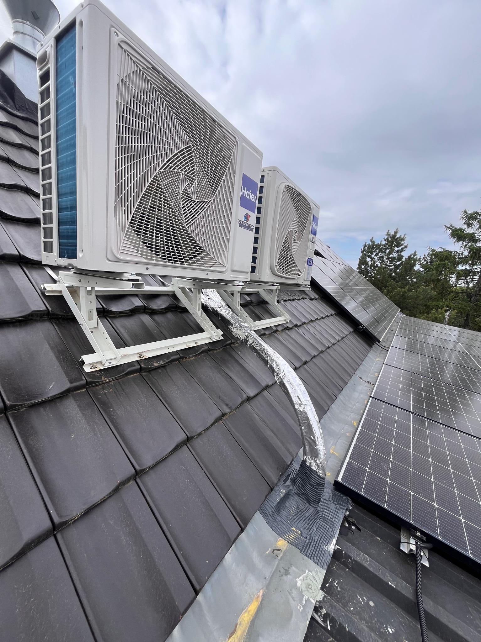 Dwa białe, zewnętrzne moduły klimatyzacji marki Haier zamontowane na metalowym stelażu na ciemnym, skośnym dachu z dachówki ceramicznej, obok paneli fotowoltaicznych, z widocznymi przewodami...