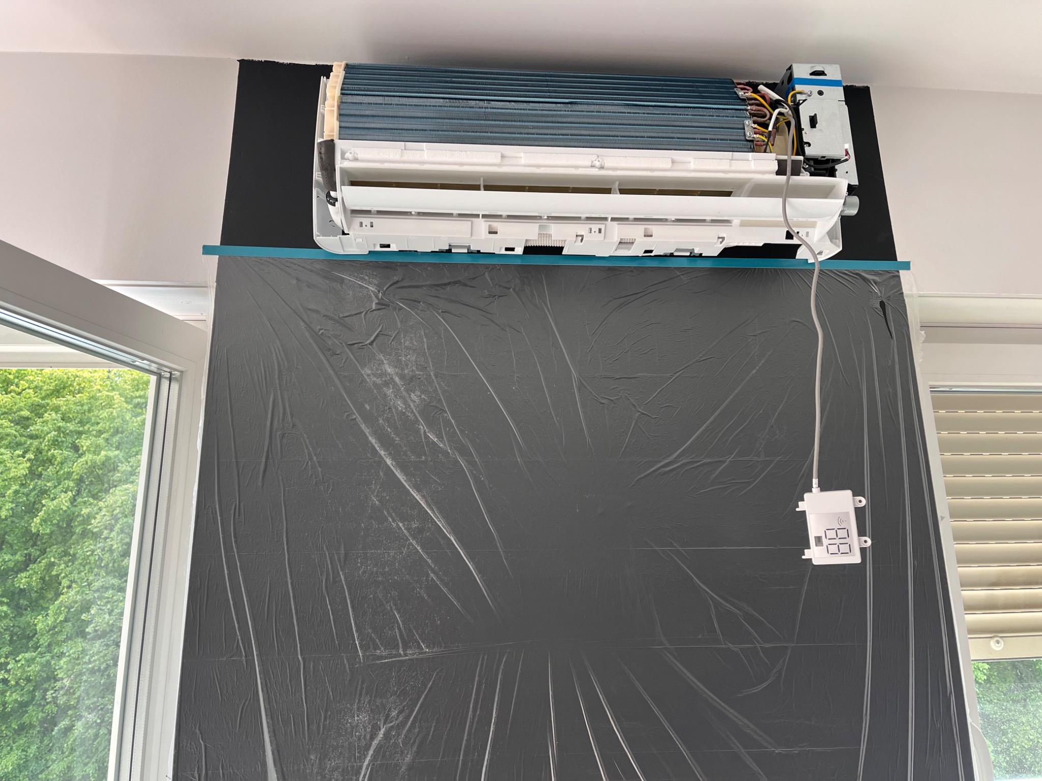 Rozłożona klimatyzacja ścienna z widocznymi elementami wewnętrznymi, zabezpieczona folią ochronną podczas serwisu, z panelem sterowania obok okna.