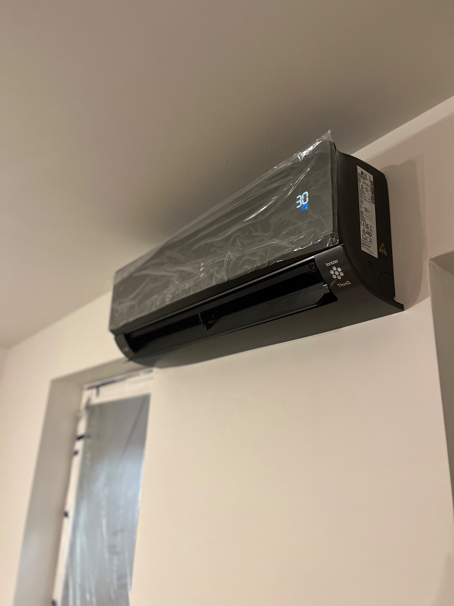 Czarna jednostka klimatyzacji LG Dual Inverter, z wyświetlaczem temperatury 30 stopni Celsjusza, zamontowana na białej ścianie, nadal zabezpieczona folią ochronną po instalacji, widoczny fragment...