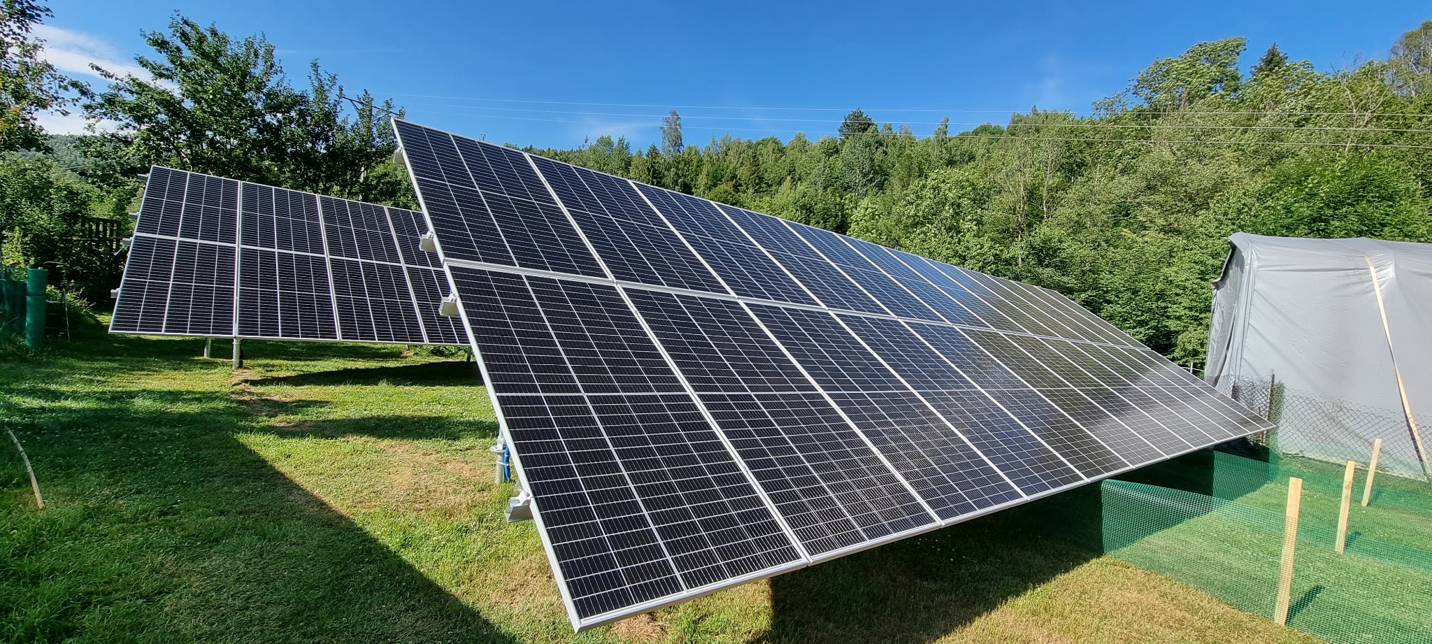 Dwie instalacje paneli słonecznych na trawniku, zalesione wzgórze w tle, zabezpieczenie z siatki wokół jednej z instalacji.