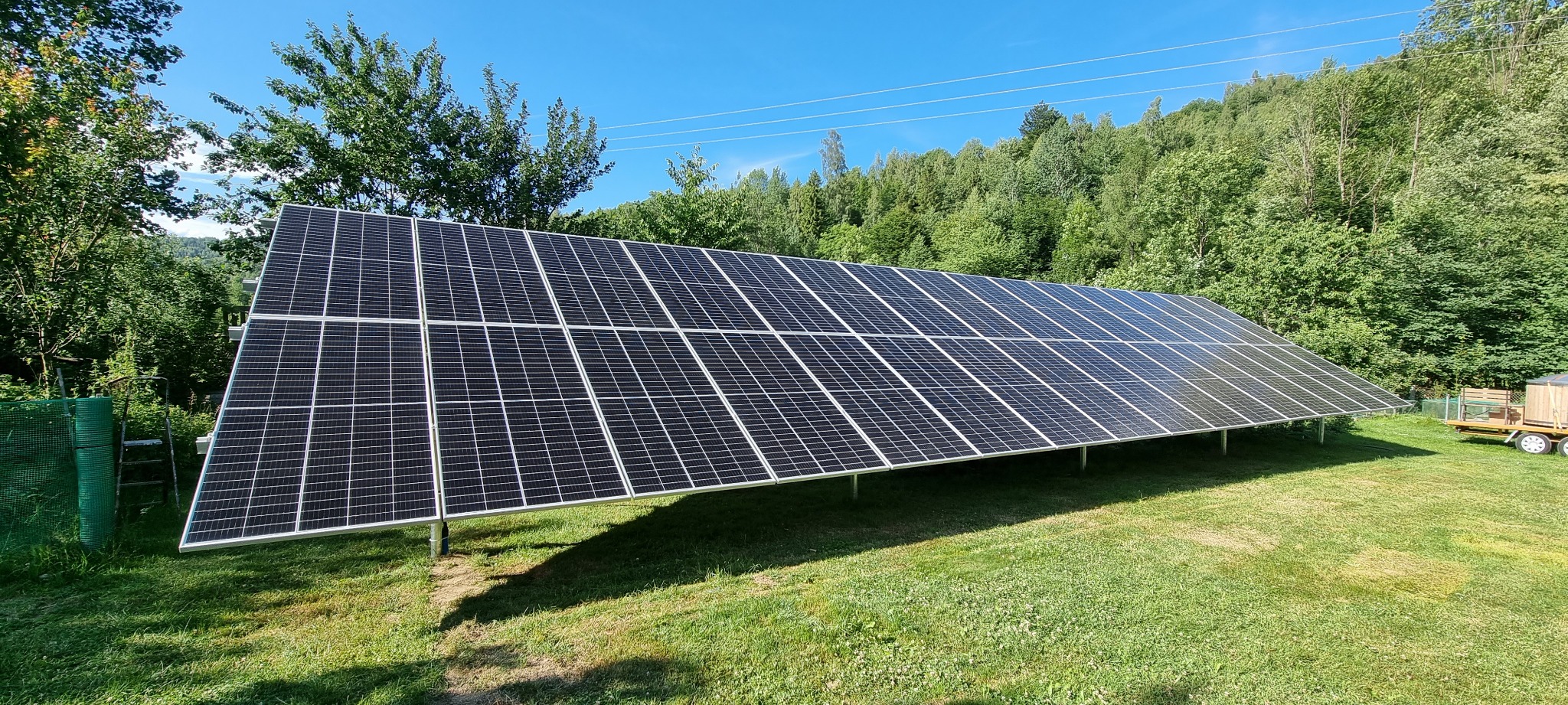 Rozległa instalacja fotowoltaiczna na metalowej konstrukcji, umieszczona na trawniku z widokiem na zalesione wzgórze w słoneczny dzień.