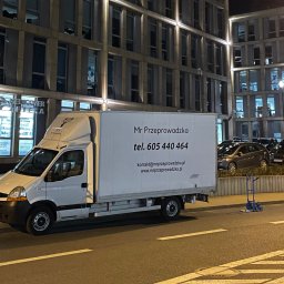 BAMI Group Sp. z o.o. - Biały samochód dostawczy firmy Mr Przeprowadzka zaparkowany na ulicy w nocy, z widoczną reklamą firmy na boku i wózkiem transportowym w tle.