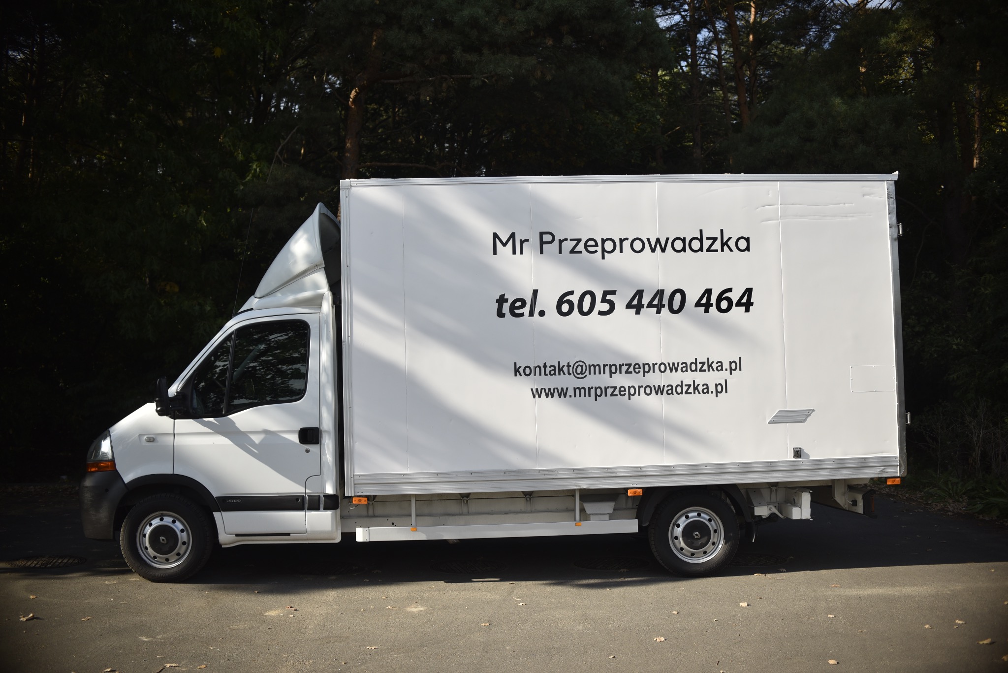 Biały samochód dostawczy z nadrukowanym napisem 'Mr Przeprowadzka', numerem telefonu i adresem strony internetowej, zaparkowany na asfalcie z drzewami w tle.