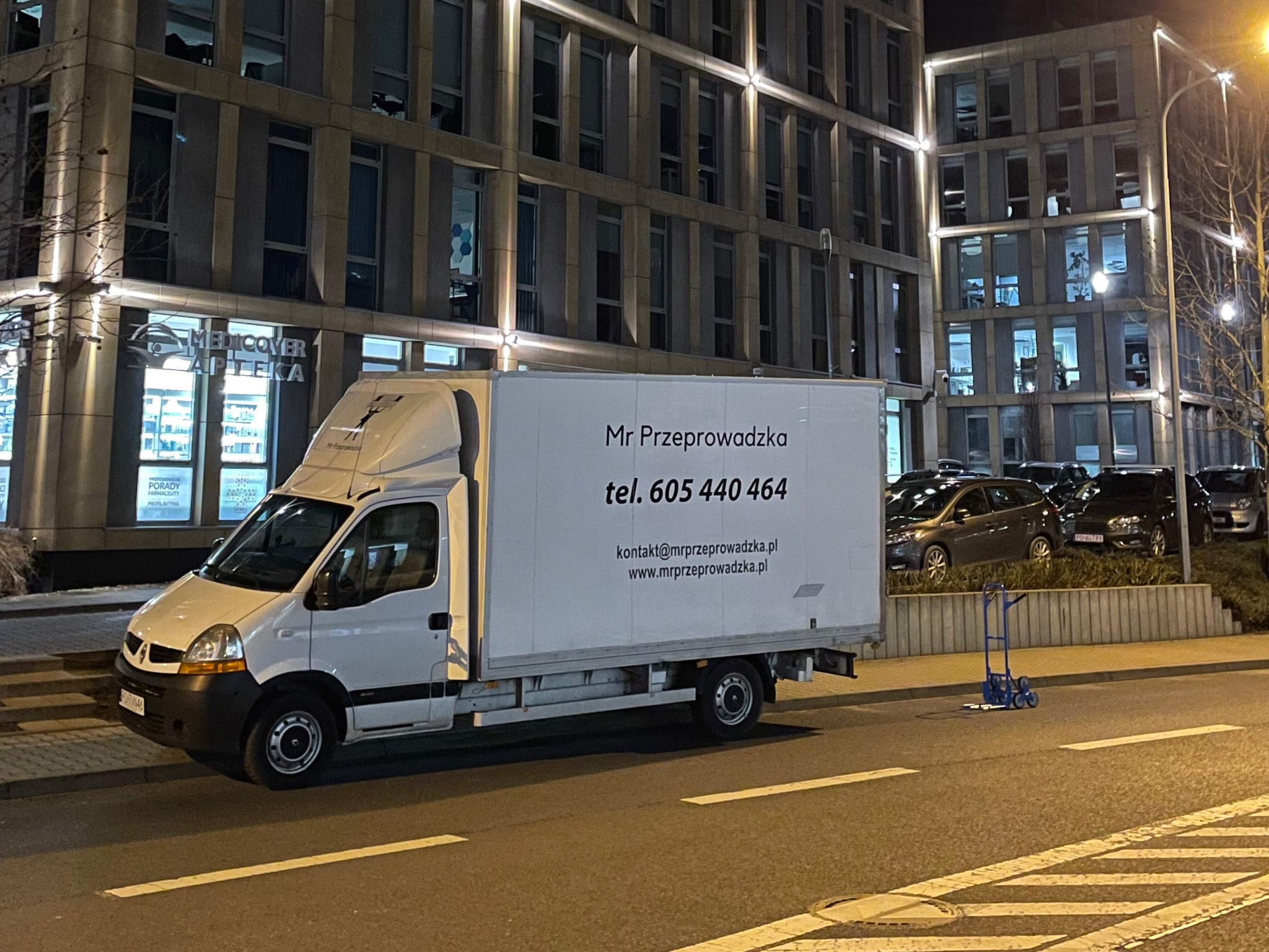 Biały samochód dostawczy firmy Mr Przeprowadzka zaparkowany na ulicy w nocy, z widoczną reklamą firmy na boku i wózkiem transportowym w tle.