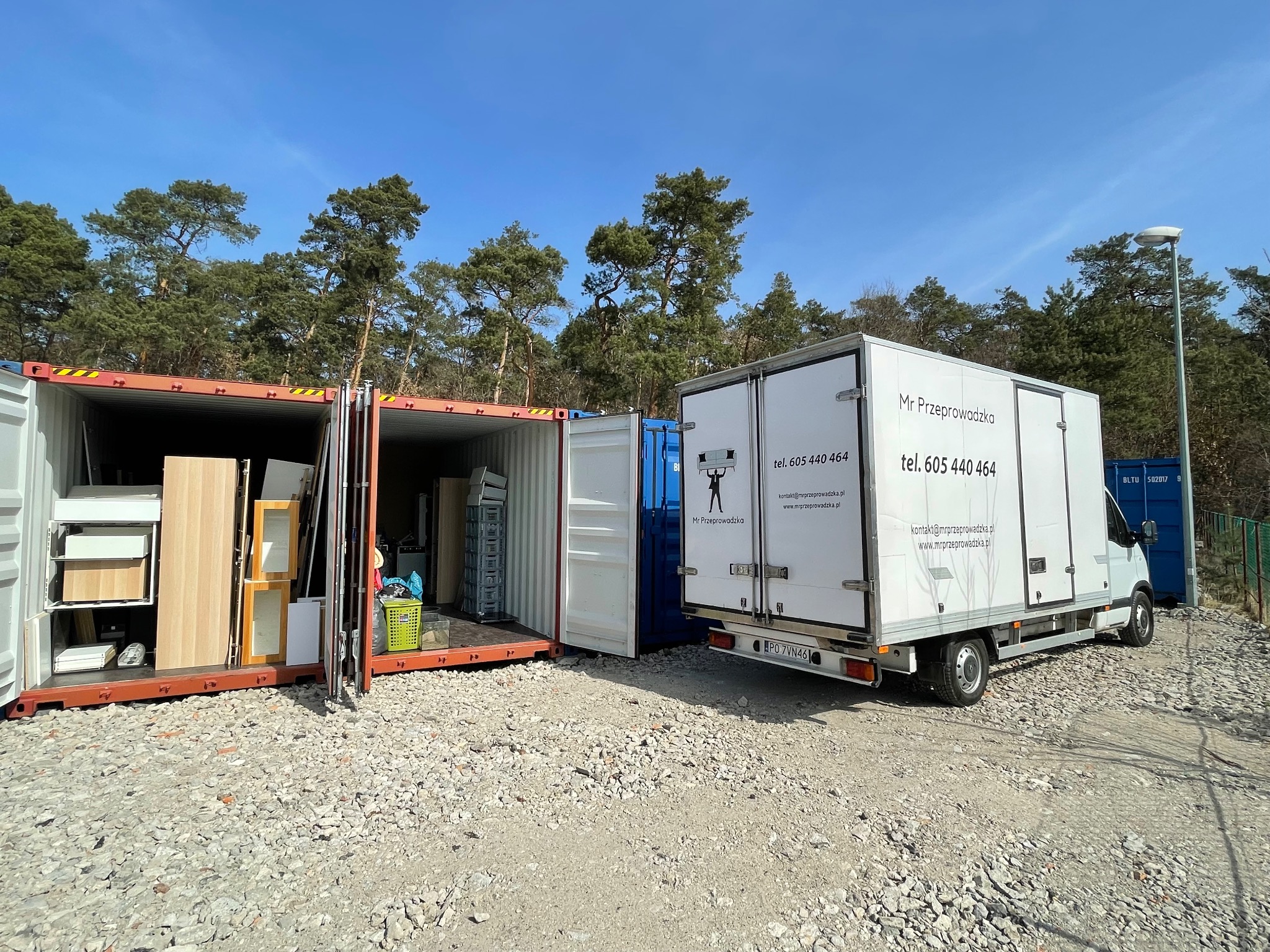 Kontenery załadowane meblami i wyposażeniem obok białego busa z logo firmy przeprowadzkowej na żwirowym placu, w tle las.