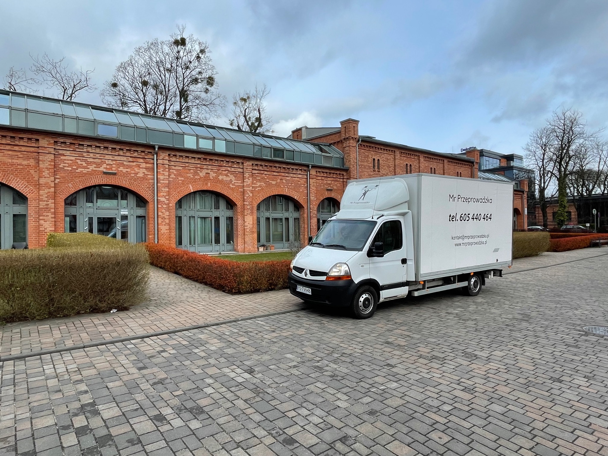 Biała furgonetka z logo firmy przeprowadzkowej zaparkowana przed budynkiem z czerwonej cegły z przeszklonym dachem, widoczne krzewy i kostka brukowa