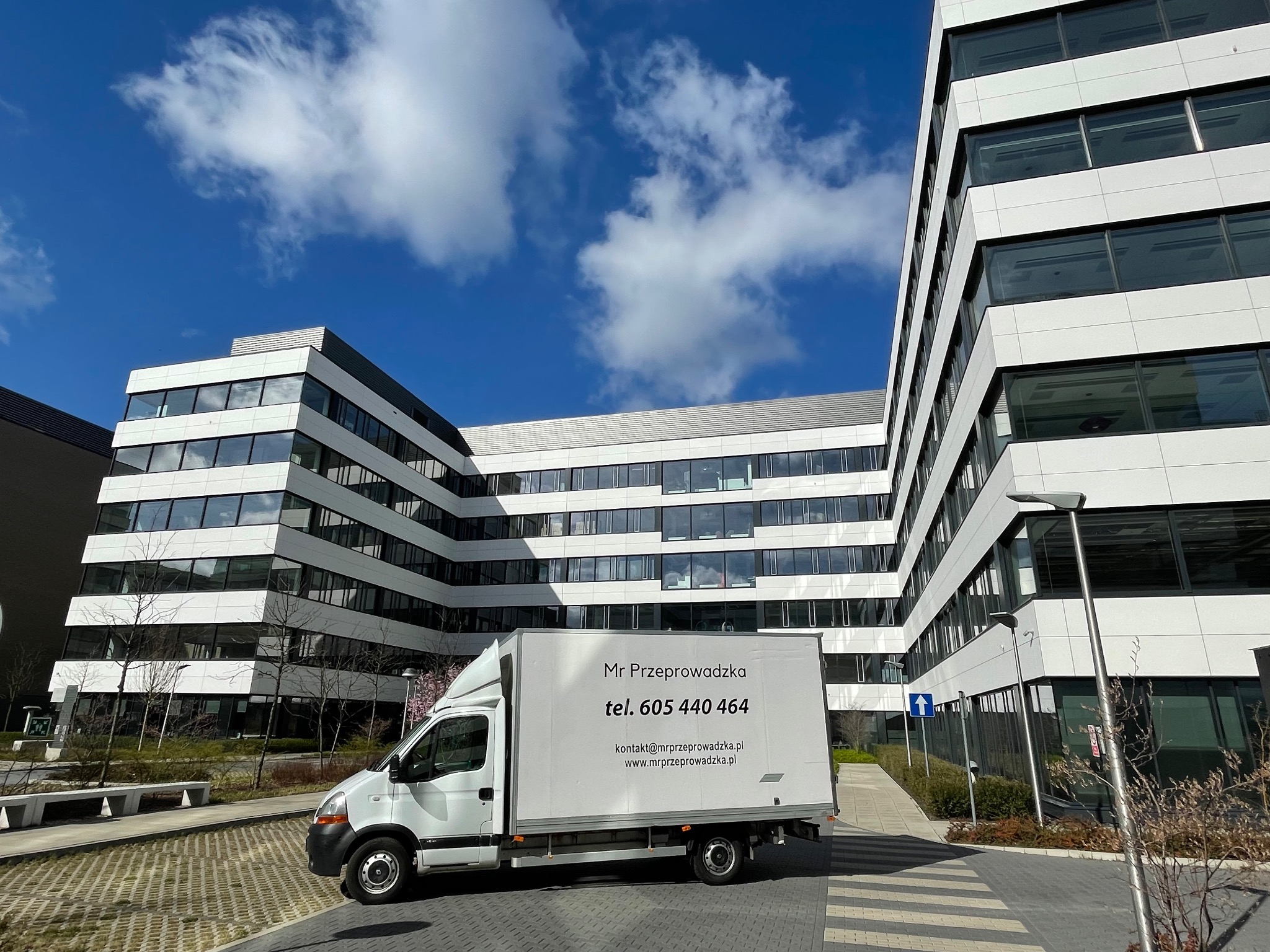 Biały bus z napisem 'Mr Przeprowadzka' na tle nowoczesnego biurowca i błękitnego nieba z chmurami.
