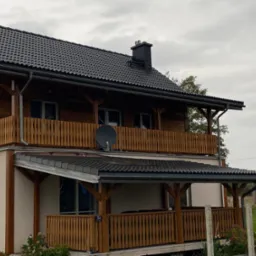 Dwupiętrowy dom z drewnianymi balkonami i nowym, ciemnoszarym dachem z blachy trapezowej oraz ciemnym kominem, widoczna antena satelitarna na balkonie.