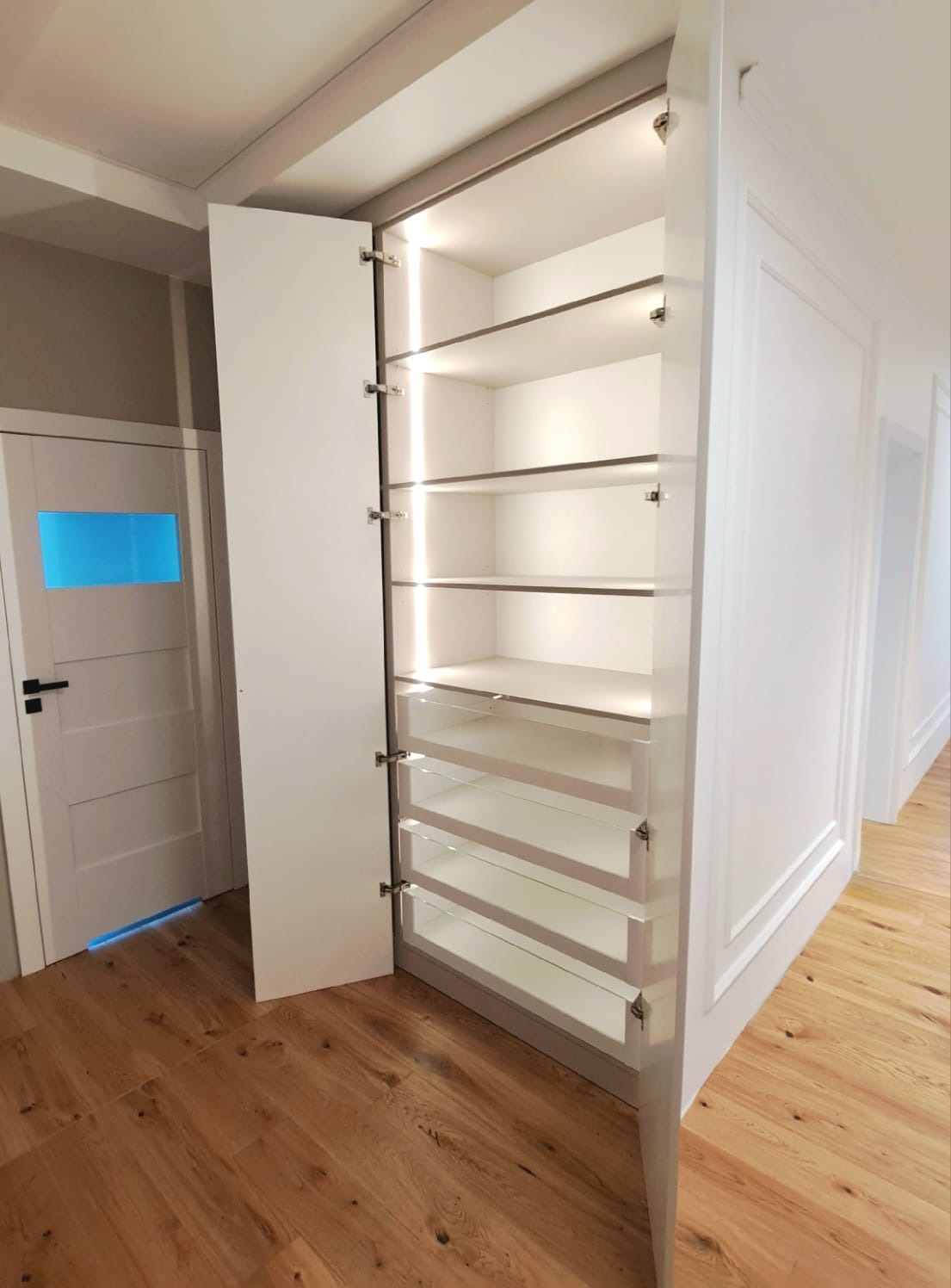 Biała szafa wnękowa z otwartymi drzwiami, podświetlonymi półkami i szufladami z przezroczystymi frontami, widoczna na tle drewnianej podłogi i białych ścian z dekoracyjnymi listwami.