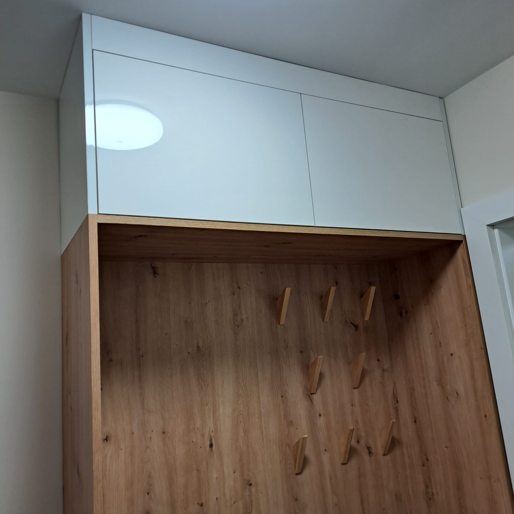 Wnęka w przedpokoju z drewnianą boazerią i wieszakami oraz białą szafką pod sufitem, oświetlenie sufitowe odbija się w lakierowanych frontach szafki.