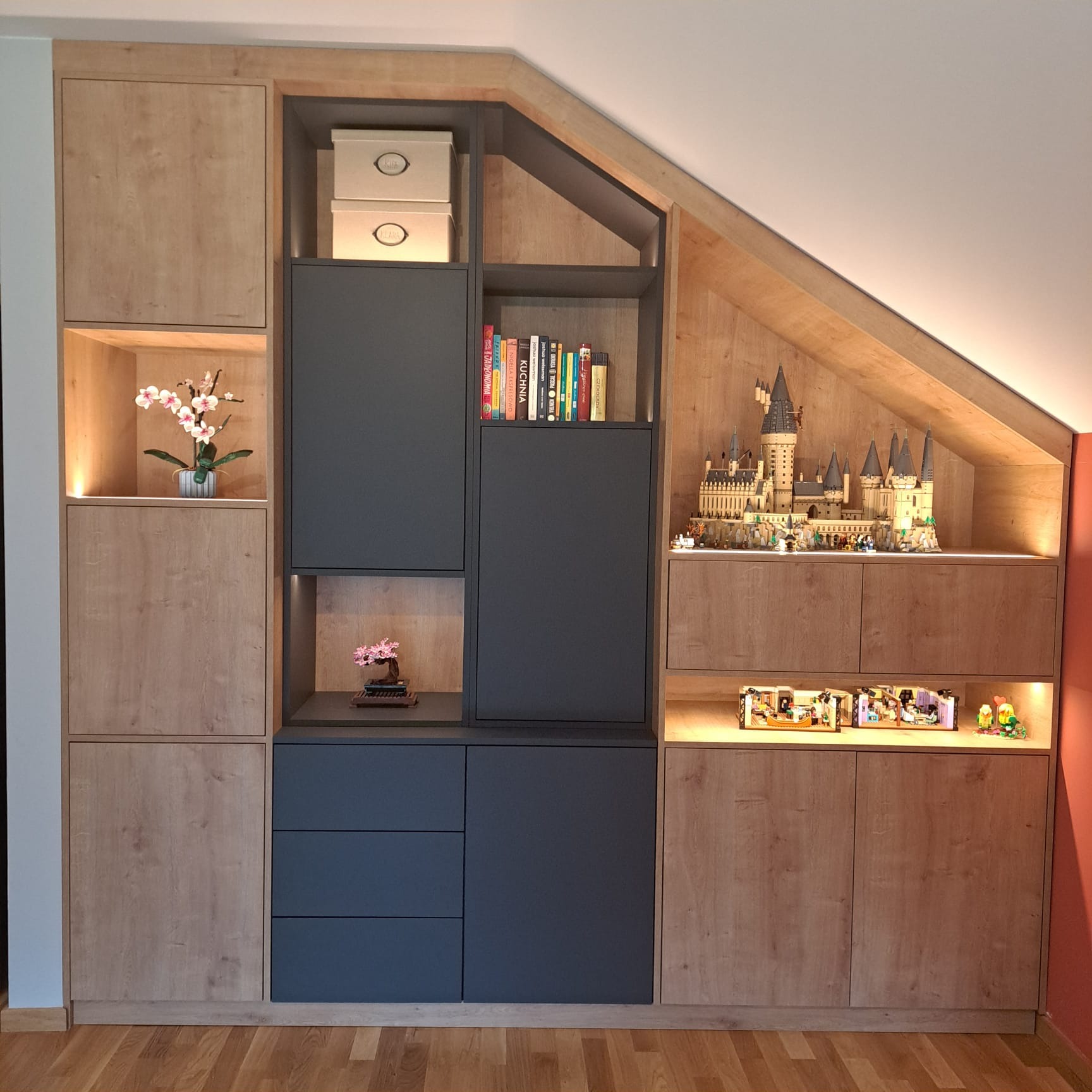 Zabudowa meblowa na wymiar, z oświetleniem LED, wkomponowana w skos dachu, z półkami na książki, dekoracje i model zamku Hogwart z klocków Lego.