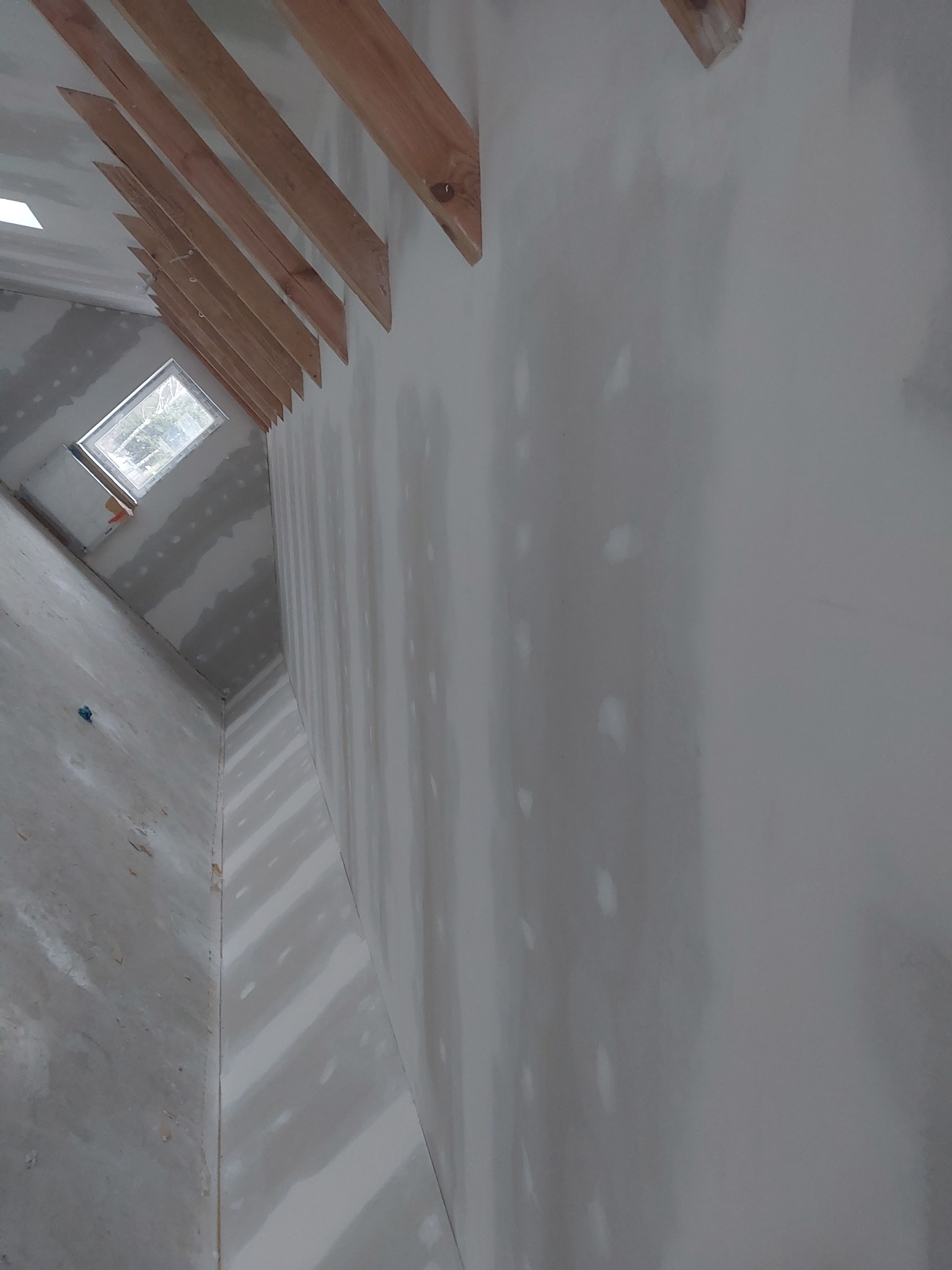 Wnętrze poddasza w trakcie prac wykończeniowych: ściany pokryte gładzią szpachlową, widoczne drewniane elementy konstrukcyjne dachu i okno dachowe.