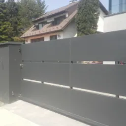 Nowoczesna, przesuwna brama w kolorze grafitowym z poziomymi prześwitami, zamontowana przed domem z brązowym dachem i jasną elewacją.